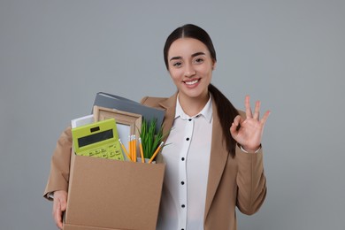 Photo of Happy unemployed woman with box of personal office belongings showing OK gesture on grey background