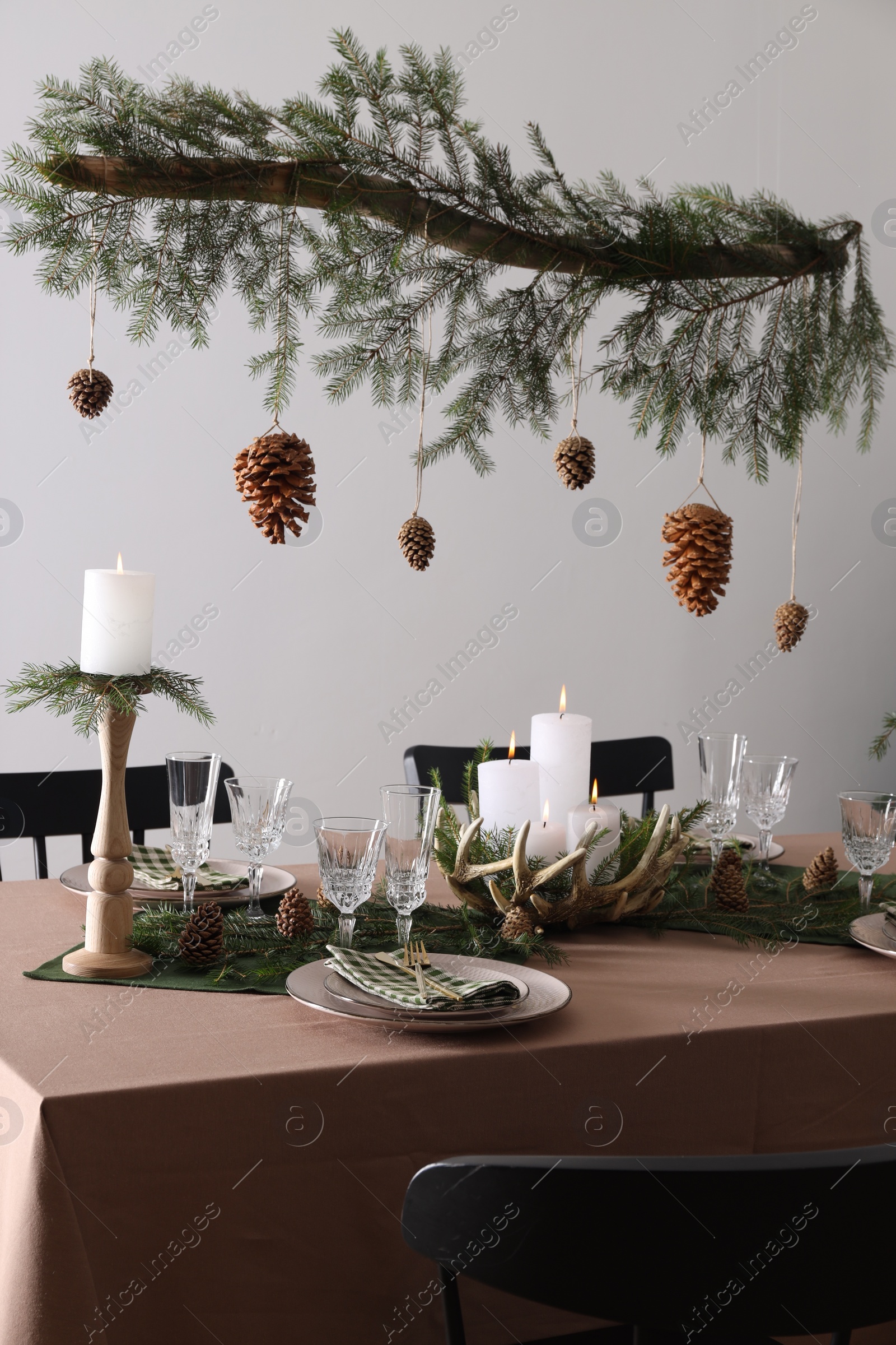 Photo of Christmas celebration. Cones hanging from fir tree branch over table with burning candles and tableware