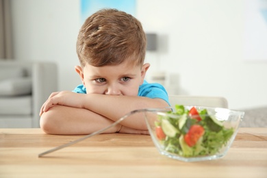 Sad little boy with bowl of vegetable salad at table in room