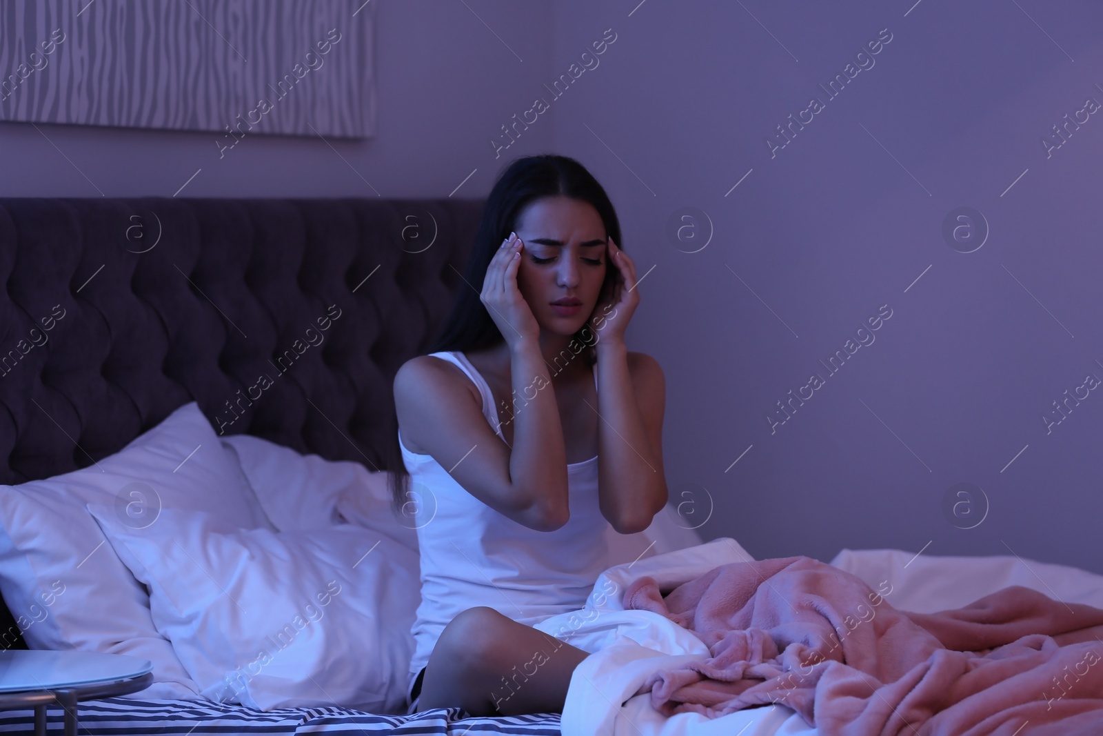 Photo of Young woman suffering from headache while sitting on bed at night. Sleeping problems