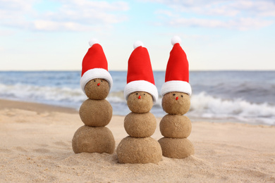 Photo of Snowmen made of sand with Santa hats on beach near sea. Christmas vacation