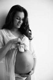 Photo of Young pregnant woman in lace nightgown holding baby shoes on light background, black and white effect. Space for text