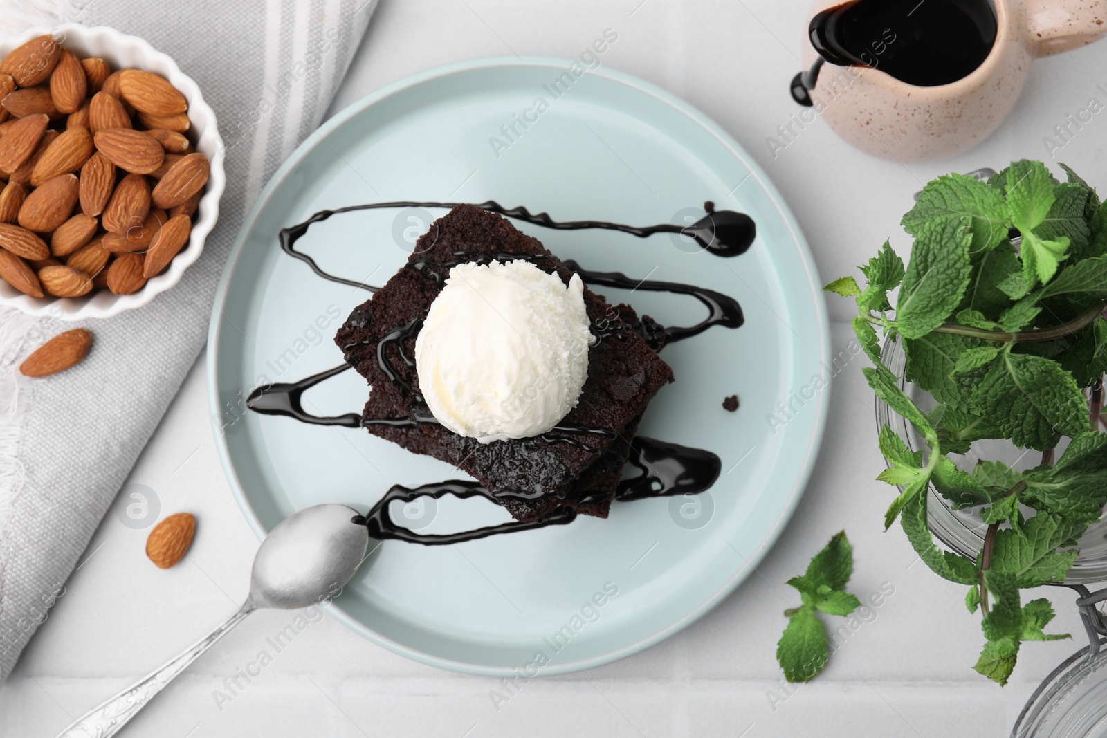 Photo of Tasty brownies served with ice cream and chocolate sauce on white tiled table, flat lay