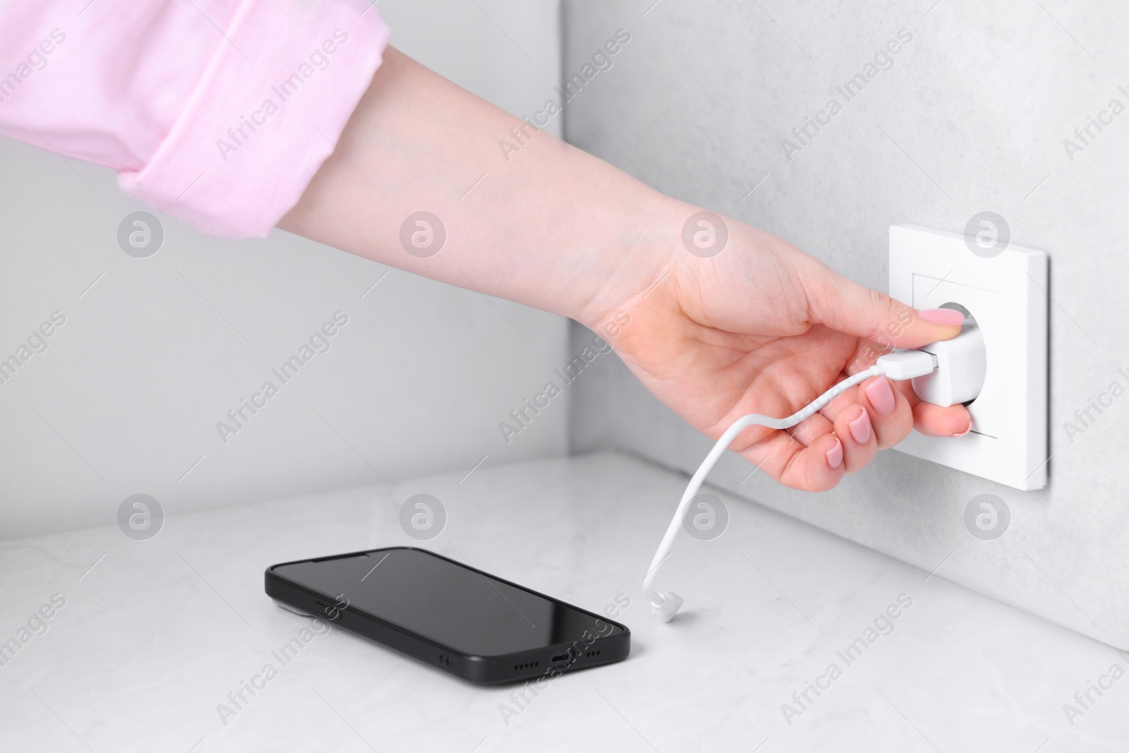 Photo of Woman plugging smartphone into power socket at white table indoors, closeup