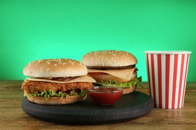 Delicious burgers and drink on wooden table against green background. Fast food menu