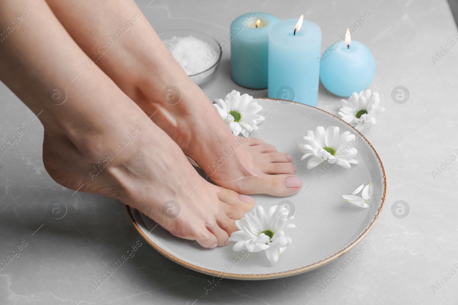 Photo of Woman soaking her feet in bowl with water and flowers on grey marble floor, closeup. Pedicure procedure
