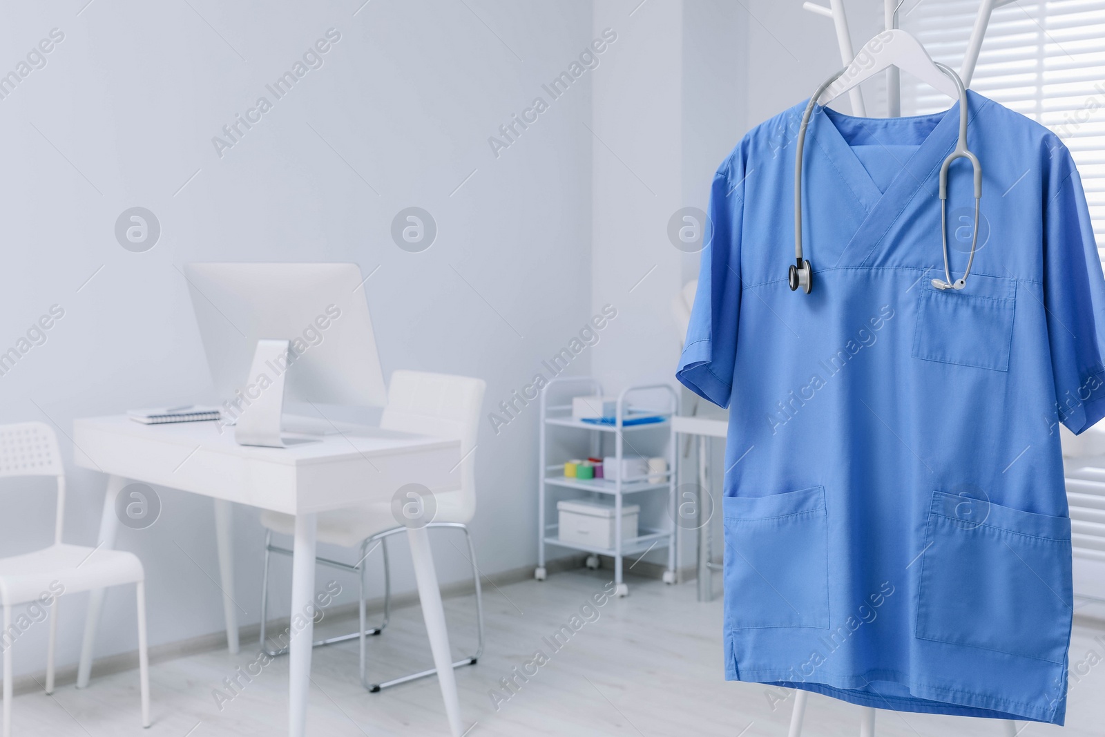 Photo of Blue medical uniform and stethoscope hanging on rack in clinic. Space for text