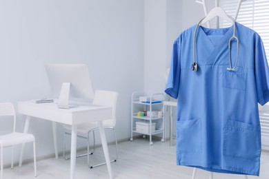Blue medical uniform and stethoscope hanging on rack in clinic. Space for text