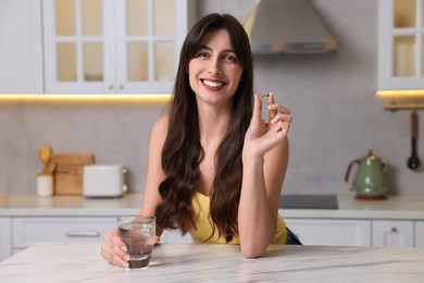 Beautiful woman with vitamin pill and glass of water at white marble table in kitchen
