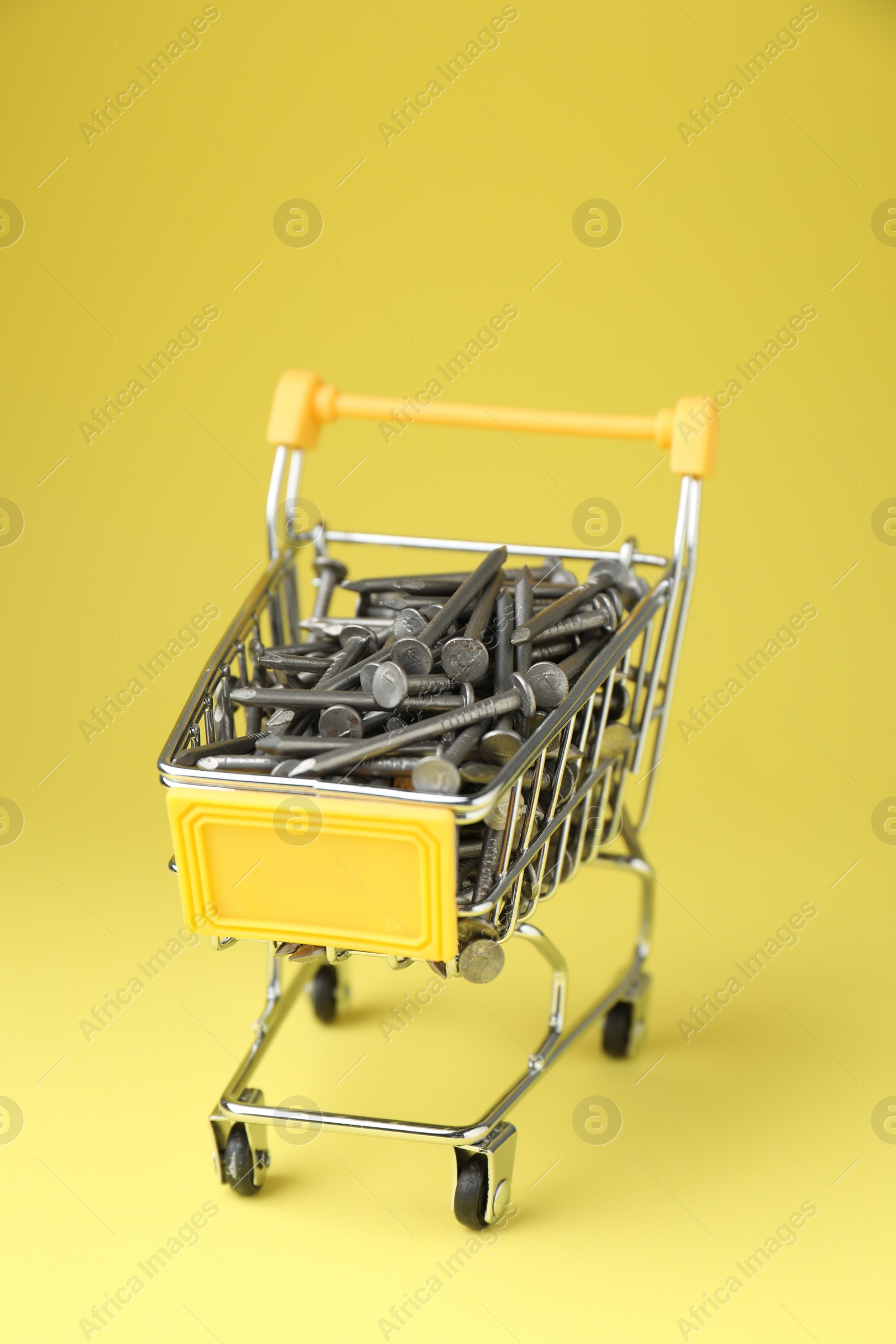 Photo of Metal nails in shopping cart on yellow background, closeup