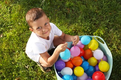 Photo of Little boy with basin of water bombs on green grass, top view