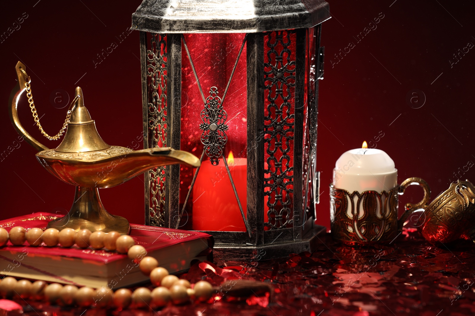 Photo of Arabic lantern, Quran, misbaha, burning candle and Aladdin magic lamp on shiny red table