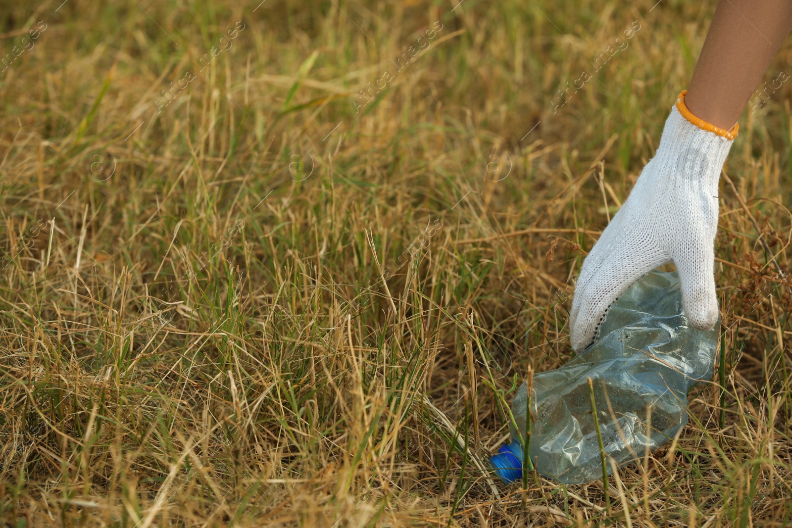 Photo of Woman in gloves picking crumpled bottle from grass outdoors, closeup. Space for text