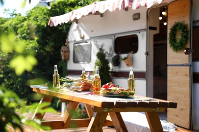 Photo of Wooden table with food and bottles of beer near motorhome on sunny day. Camping season