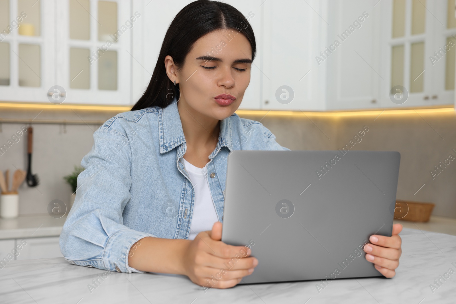 Photo of Young woman having video chat via laptop and sending air kiss at table in kitchen