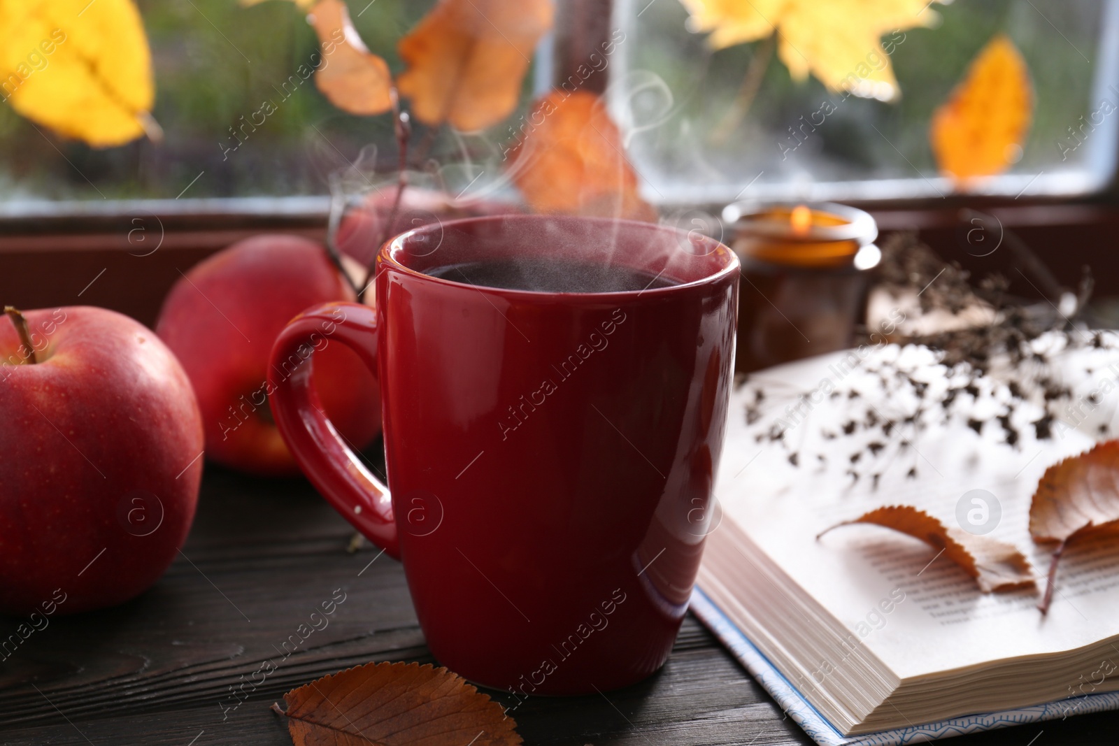 Photo of Cup of hot drink, open book and ripe apples on wooden table near window, closeup. Cozy autumn atmosphere