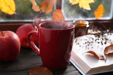 Cup of hot drink, open book and ripe apples on wooden table near window, closeup. Cozy autumn atmosphere