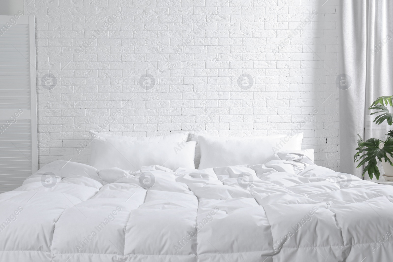 Photo of Large comfortable bed with pillows and blanket near white brick wall indoors. Stylish interior