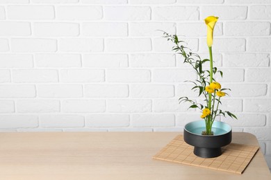 Photo of Beautiful ikebana for stylish house decor. Floral composition with fresh calla, chrysanthemum flowers and branches on wooden table near white brick wall, space for text