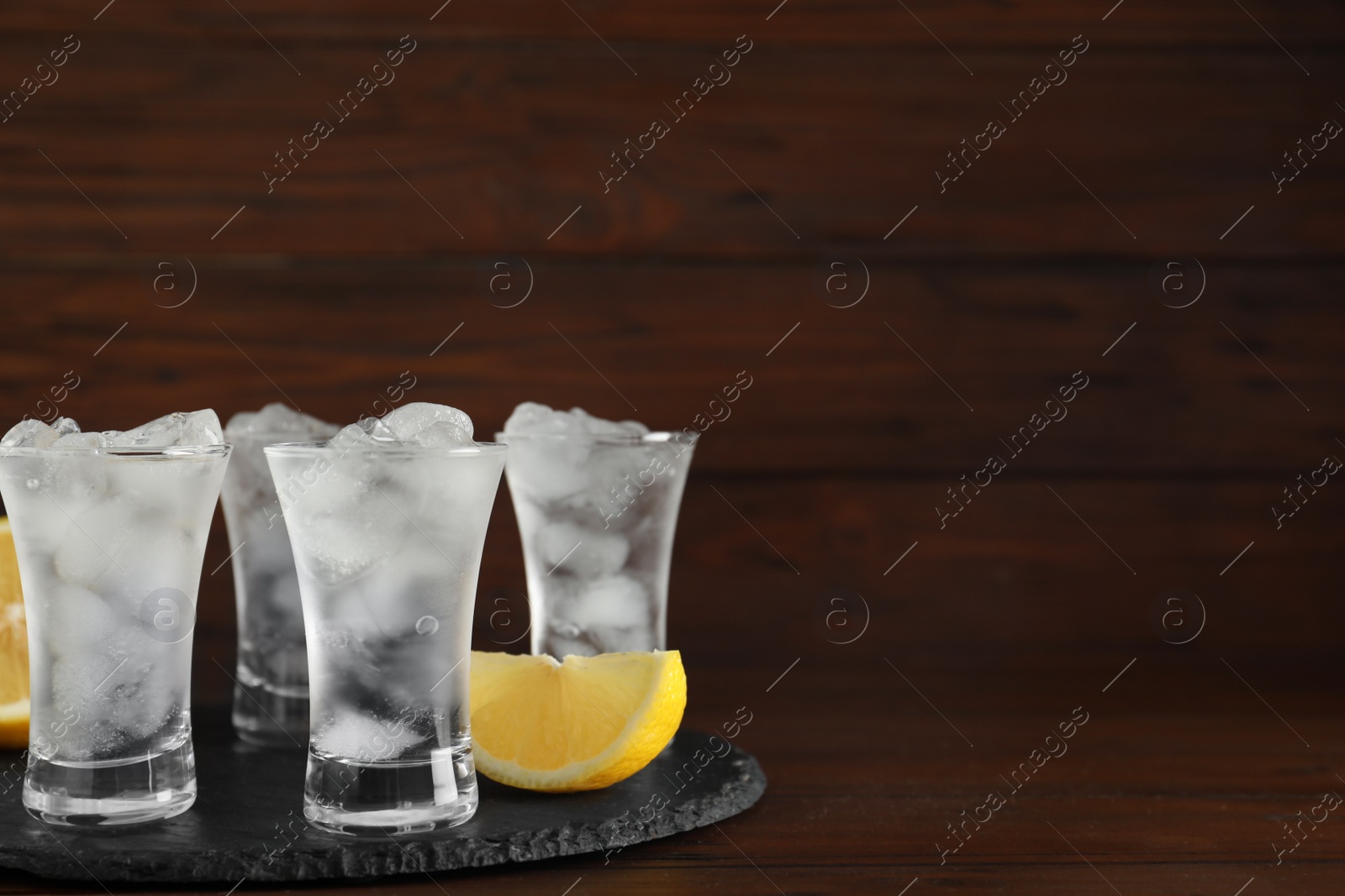 Photo of Shot glasses with vodka, ice and lemon on table against wooden background. Space for text