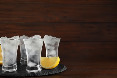 Shot glasses with vodka, ice and lemon on table against wooden background. Space for text