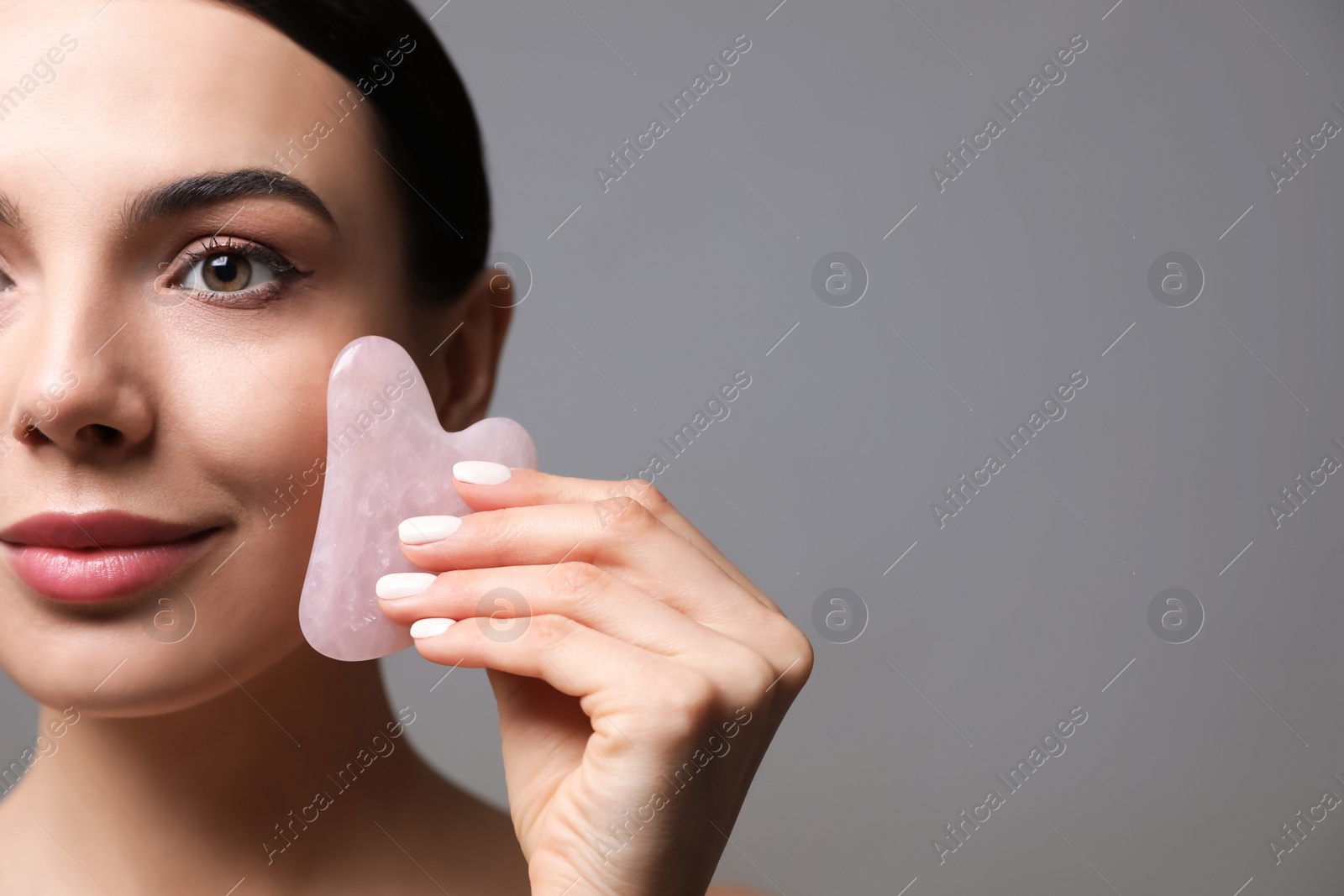 Photo of Beautiful young woman doing facial massage with gua sha tool on grey background, closeup. Space for text