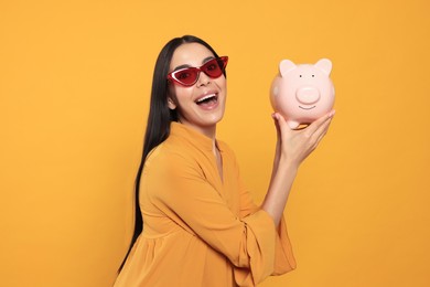 Emotional young woman with piggy bank on orange background