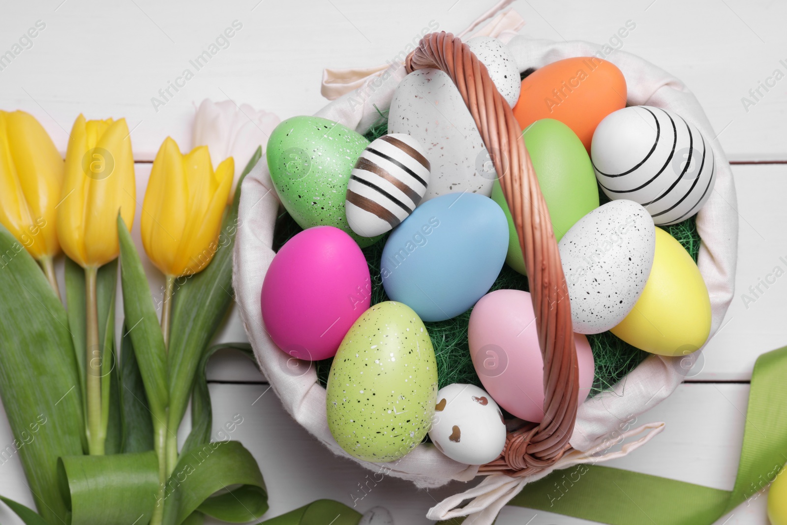 Photo of Easter eggs in basket, tulip flowers and ribbon on white wooden table, flat lay