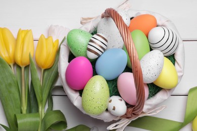 Easter eggs in basket, tulip flowers and ribbon on white wooden table, flat lay