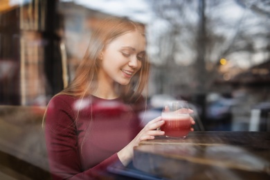 Pretty young woman with cocktail at table in cafe, view from outdoors through window