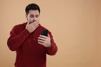 Shocked young man using smartphone on beige background, space for text