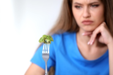Blurred view of unhappy woman against light background, focus on fork with broccoli