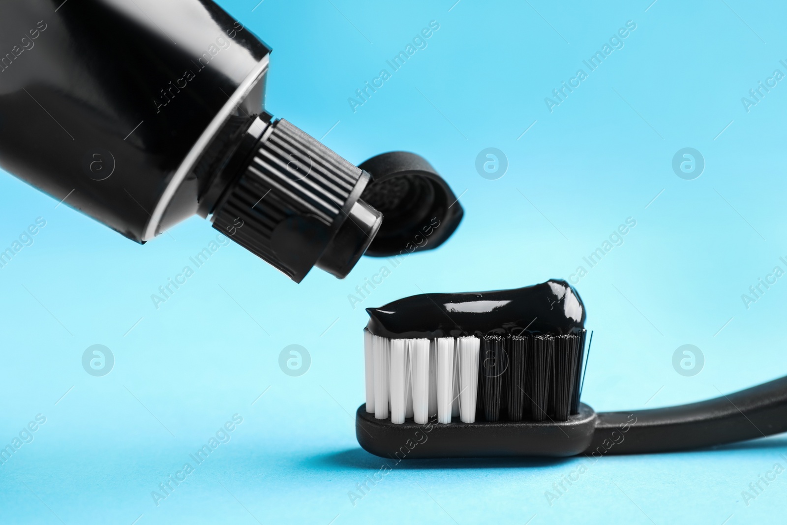 Photo of Applying charcoal toothpaste on brush against light blue background, closeup