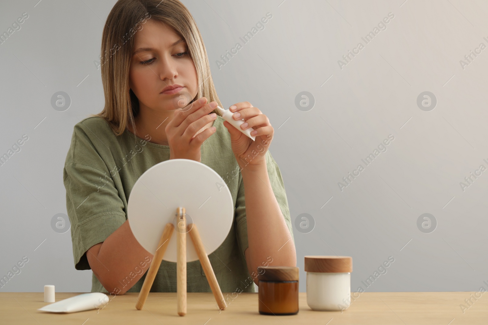 Photo of Sleep deprived young woman covering up dark circles with concealer near mirror at home