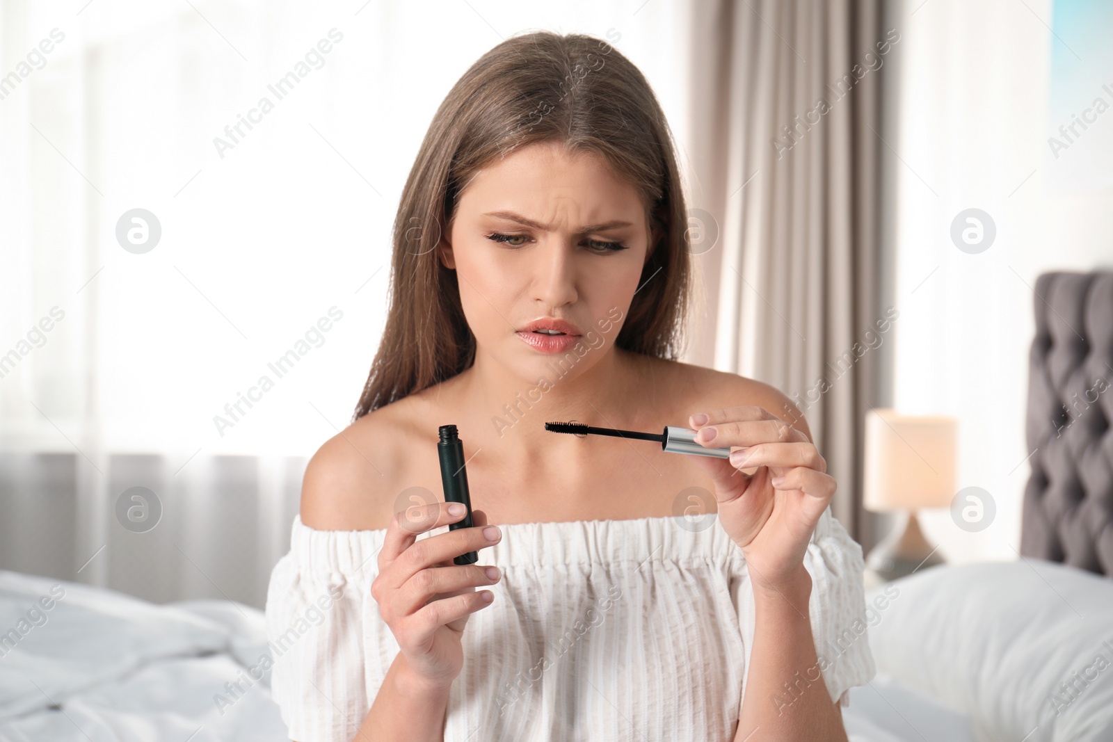 Photo of Emotional young woman holding mascara and brush with fallen eyelashes indoors