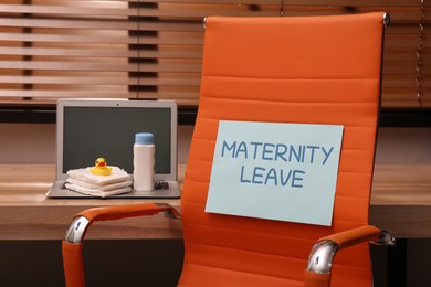 Photo of Baby accessories and laptop on wooden table near chair with note Maternity Leave in office