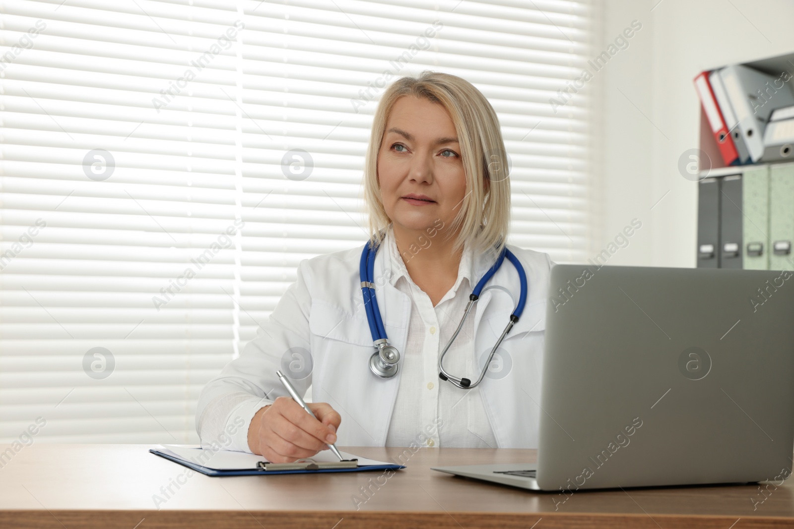 Photo of Doctor with laptop and clipboard writing at wooden table in clinic