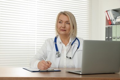 Doctor with laptop and clipboard writing at wooden table in clinic