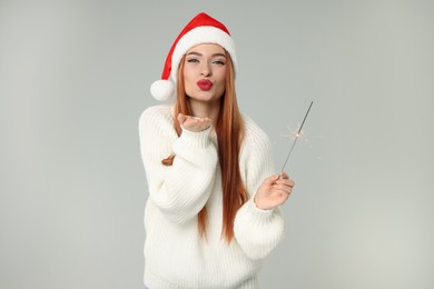 Young woman in Santa hat with burning sparkler blowing kiss on light grey background. Christmas celebration