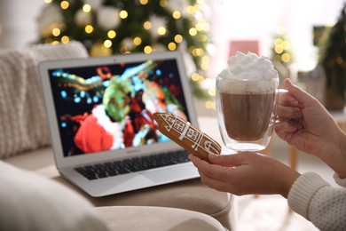 MYKOLAIV, UKRAINE - DECEMBER 25, 2020: Woman with gingerbread and cup of coffee watching The Grinch movie on laptop at home, closeup. Cozy winter holidays atmosphere