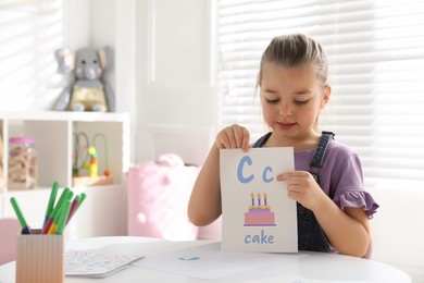 Photo of Little girl holding card with letter C and word Cake in classroom at English lesson