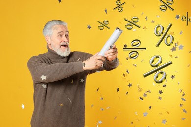 Image of Discount offer. Happy senior man blowing up party popper on golden background. Confetti and percent signs in air