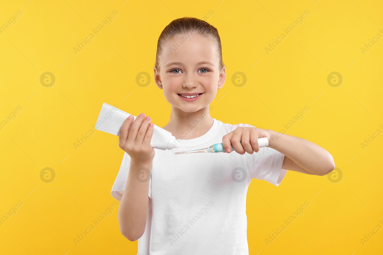 Photo of Happy girl squeezing toothpaste from tube onto electric toothbrush on yellow background