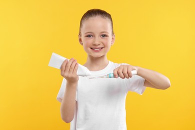 Happy girl squeezing toothpaste from tube onto electric toothbrush on yellow background