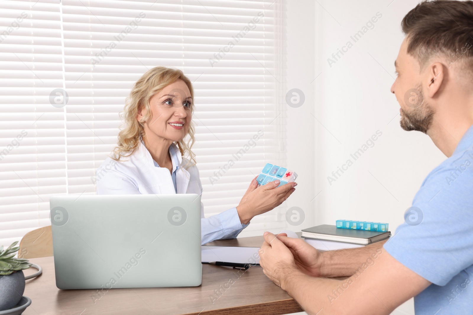 Photo of Doctor holding box of pills and consulting patient at table in clinic