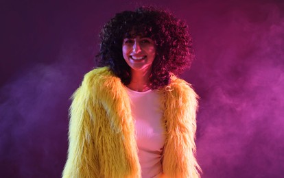 Beautiful young woman in yellow fur coat on color background in neon lights and smoke