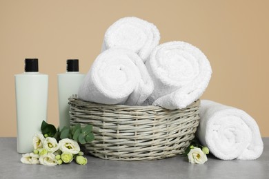Photo of Soft rolled towels in wicker basket, bottles of cosmetic products and flowers on grey table