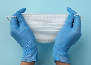 Doctor in medical gloves holding protective mask on light blue background, closeup