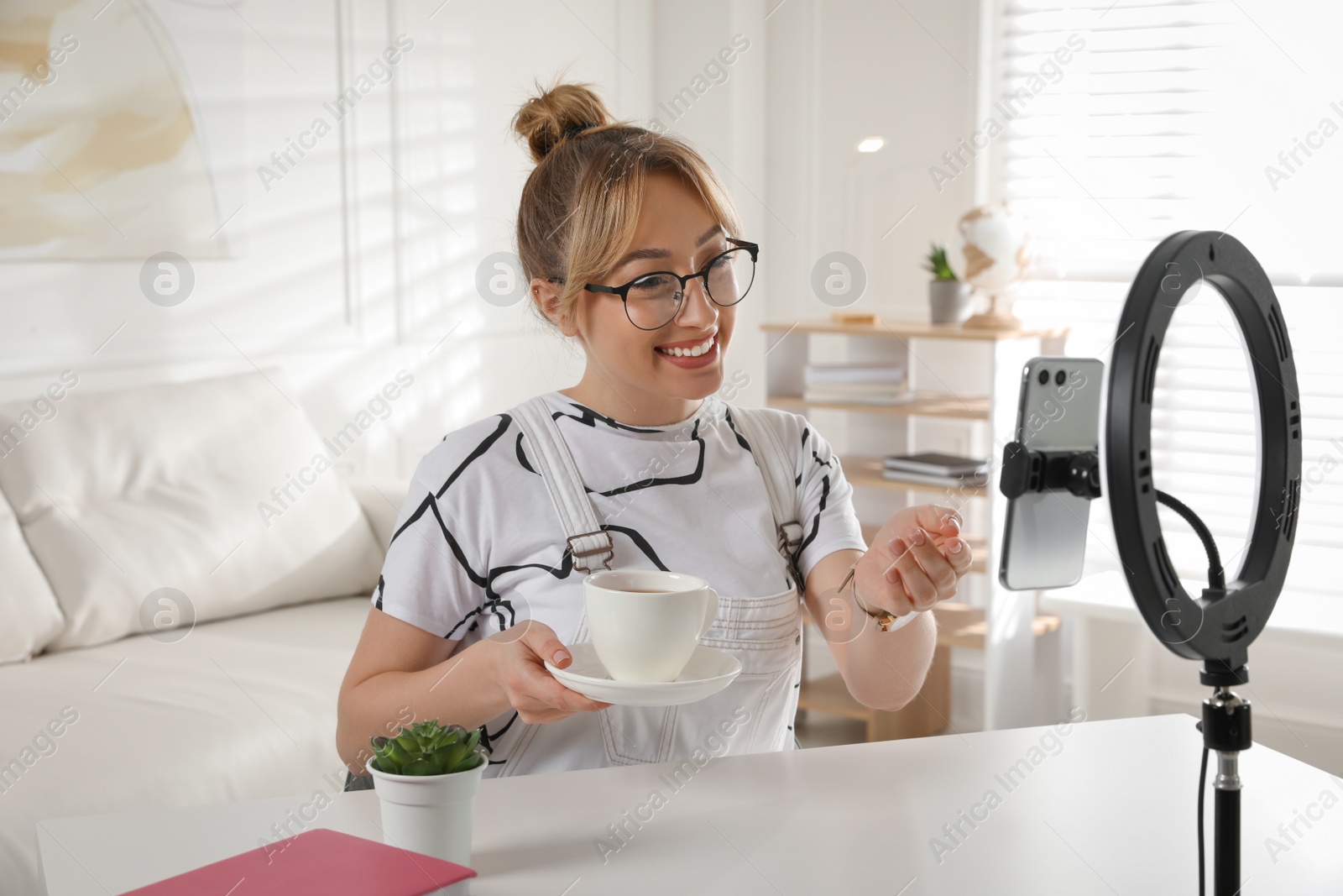 Photo of Blogger with cup of tea recording video at table indoors. Using ring lamp and smartphone