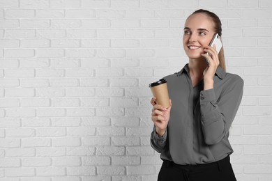Photo of Happy young secretary with paper cup of drink talking on smartphone near white brick wall, space for text